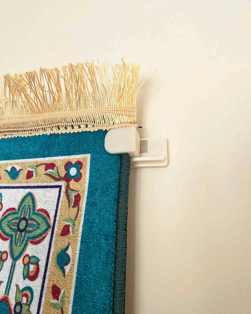 Close-up photo of a clamp at the top corner of a hung prayer mat, sitting on its wall mount just below the tassels