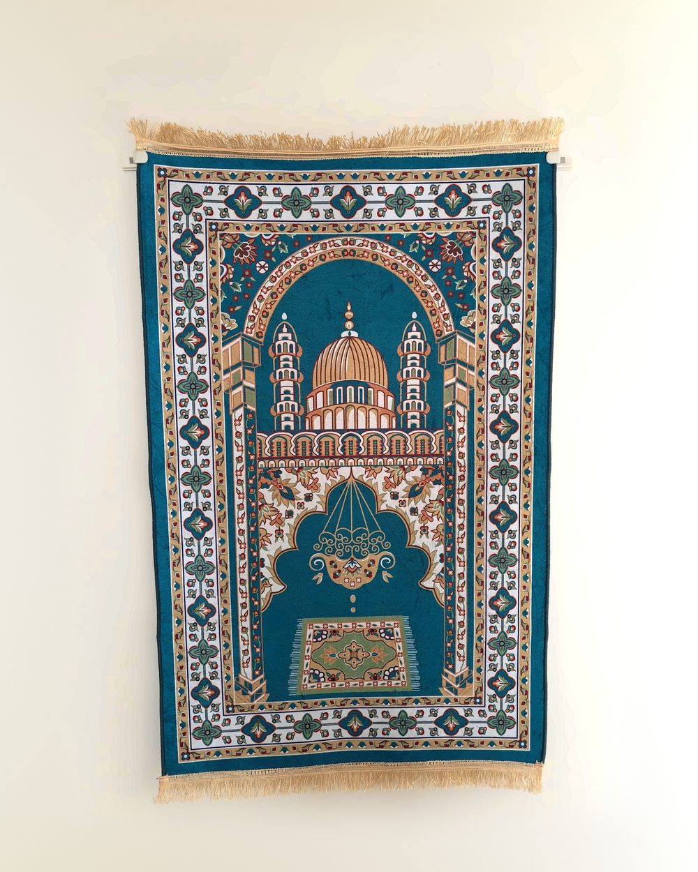 A teal and gold prayer mat hanging flat on a cream wall like a painting, held at its top corners by two small ivory clamps